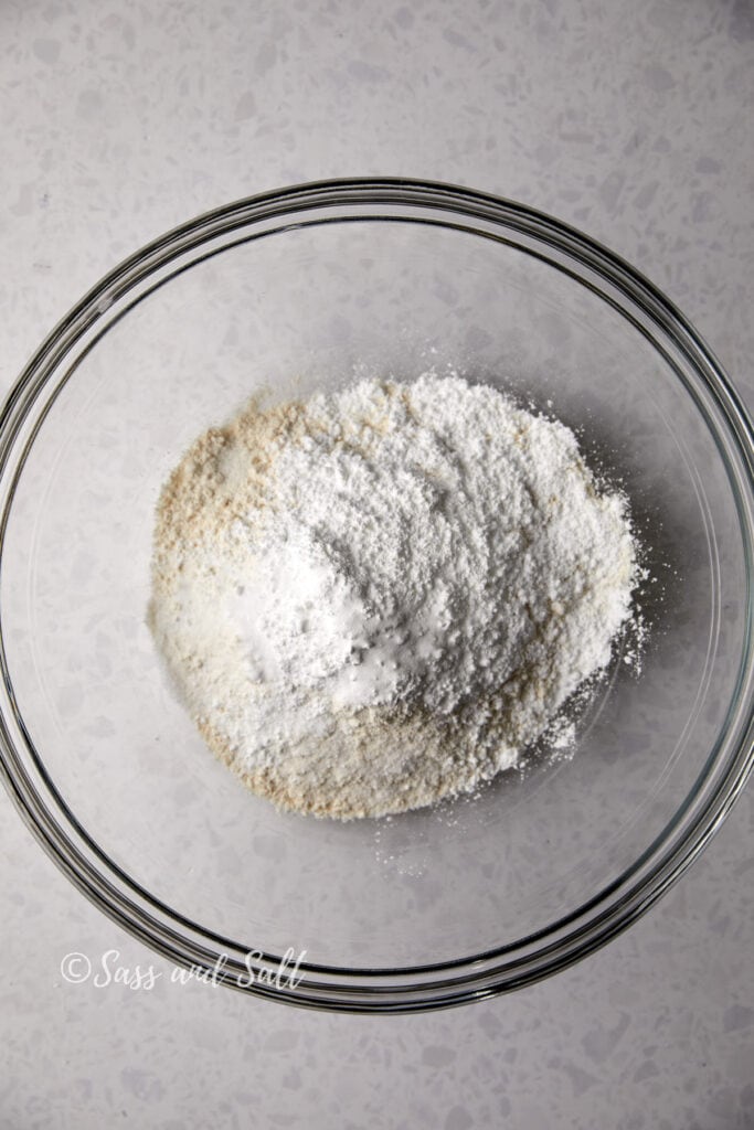 A transparent bowl containing a blend of white flour and other dry ingredients on a textured surface, seen from an overhead angle.