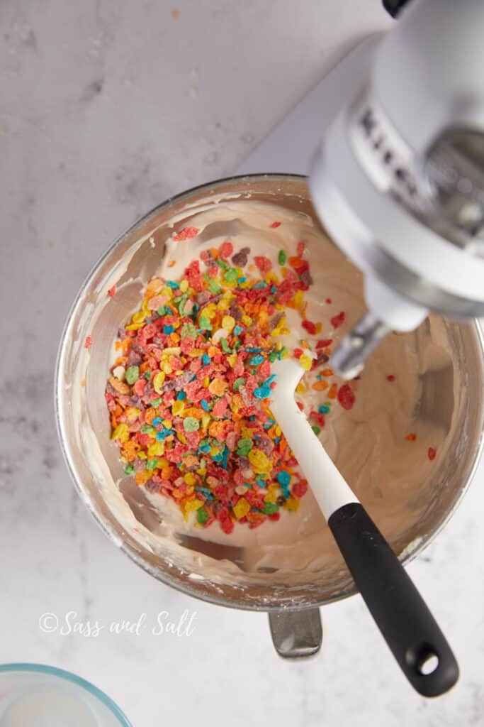 A mixer bowl with cupcake mixture, a spatula, and a vibrant sprinkle of Fruity Pebbles cereal on top, set against a marble background, with "Sass and Salt" watermark.




