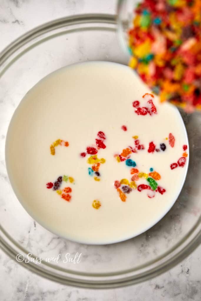 A clear glass bowl filled with whole milk with colorful Fruity Pebbles cereal being poured into it, some cereal pieces floating on the surface, set against a white marble background. 