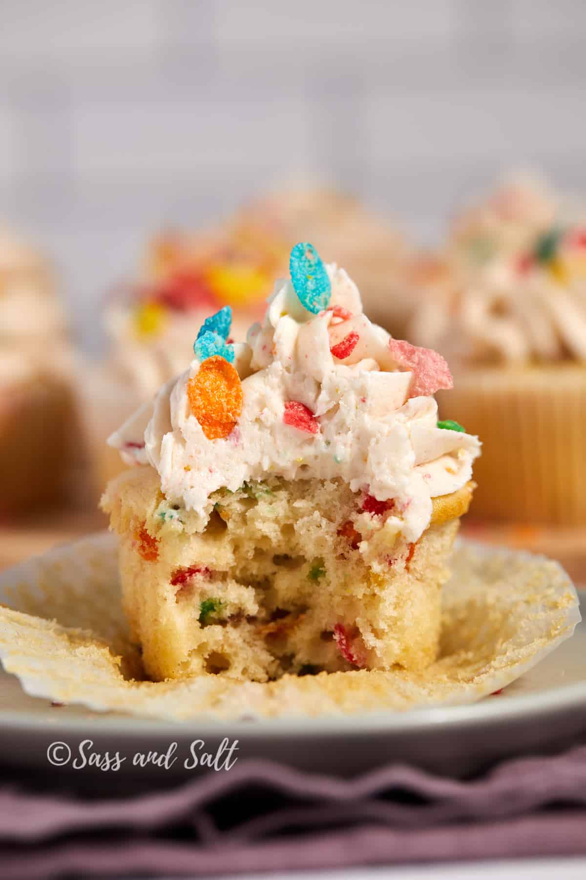 A delicious Fruity Pebbles cupcake with a bite taken out of it, revealing the colorful cereal pieces mixed into the batter. The cupcake is topped with creamy frosting sprinkled with more Fruity Pebbles, creating a playful and tempting dessert. The cupcake sits on a plate with a crumpled wrapper, and the background features more cupcakes and a soft, textured purple cloth.
