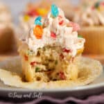 A tempting close-up of a partially eaten Fruity Pebbles cupcake, revealing colorful Fruity Pebbles inside, topped with a generous swirl of creamy Fruity Pebbles buttercream and topped with Fruity Pebbles.
