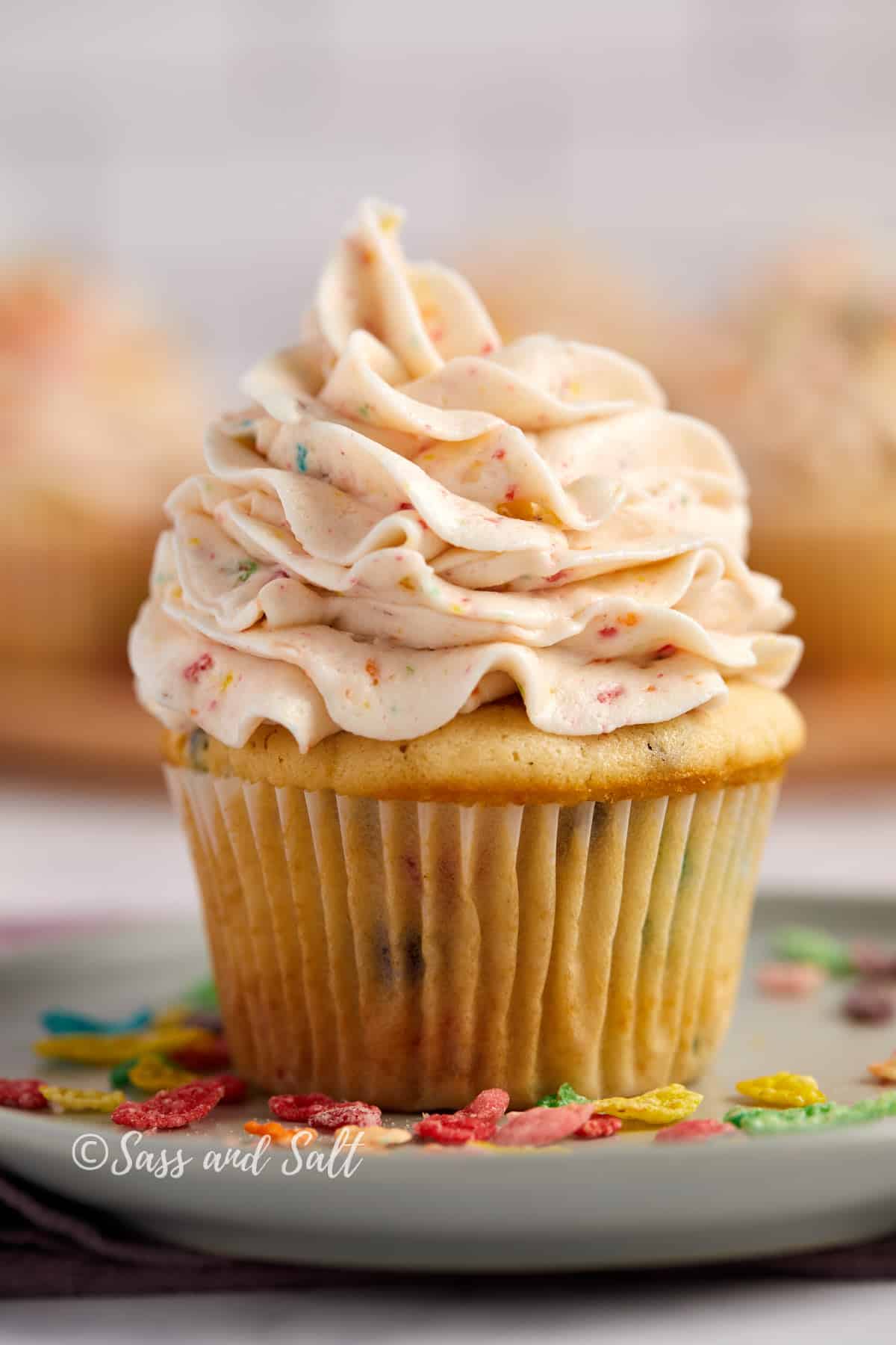 A Fruity pebbles  cupcake topped with a generous swirl of Fruity Pebbles-infused buttercream frosting, presented on a plate with Fruity Pebbles scattered around.