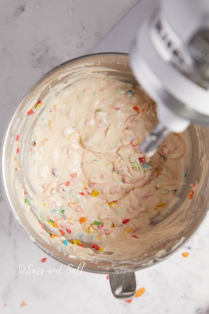 A cupcake batter with colorful Fruity Pebbles mixed in, pictured in a stainless steel mixing bowl under a mixer, displayed against a marble background with the "Sass and Salt" watermark.