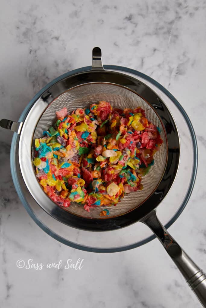 A stainless steel sieve full of colorful, wet Fruity Pebbles cereal rests on top of a glass bowl, against a marble countertop background. The vibrant cereal pieces are clumped together, suggesting they've been soaked and drained. 
