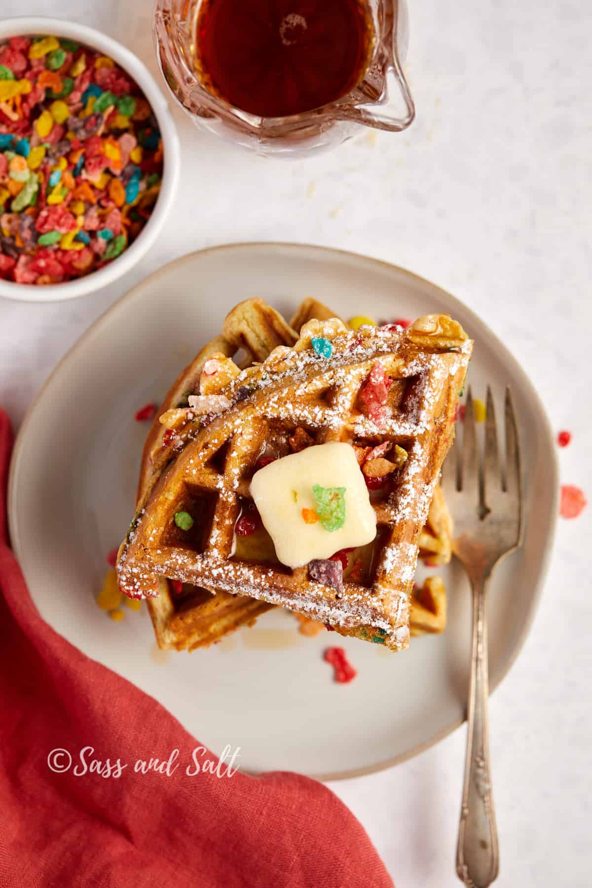 A plate of freshly-made waffles topped with a pat of butter and colorful cereal, sprinkled with powdered sugar, ready to be drizzled with syrup from the pitcher beside it.