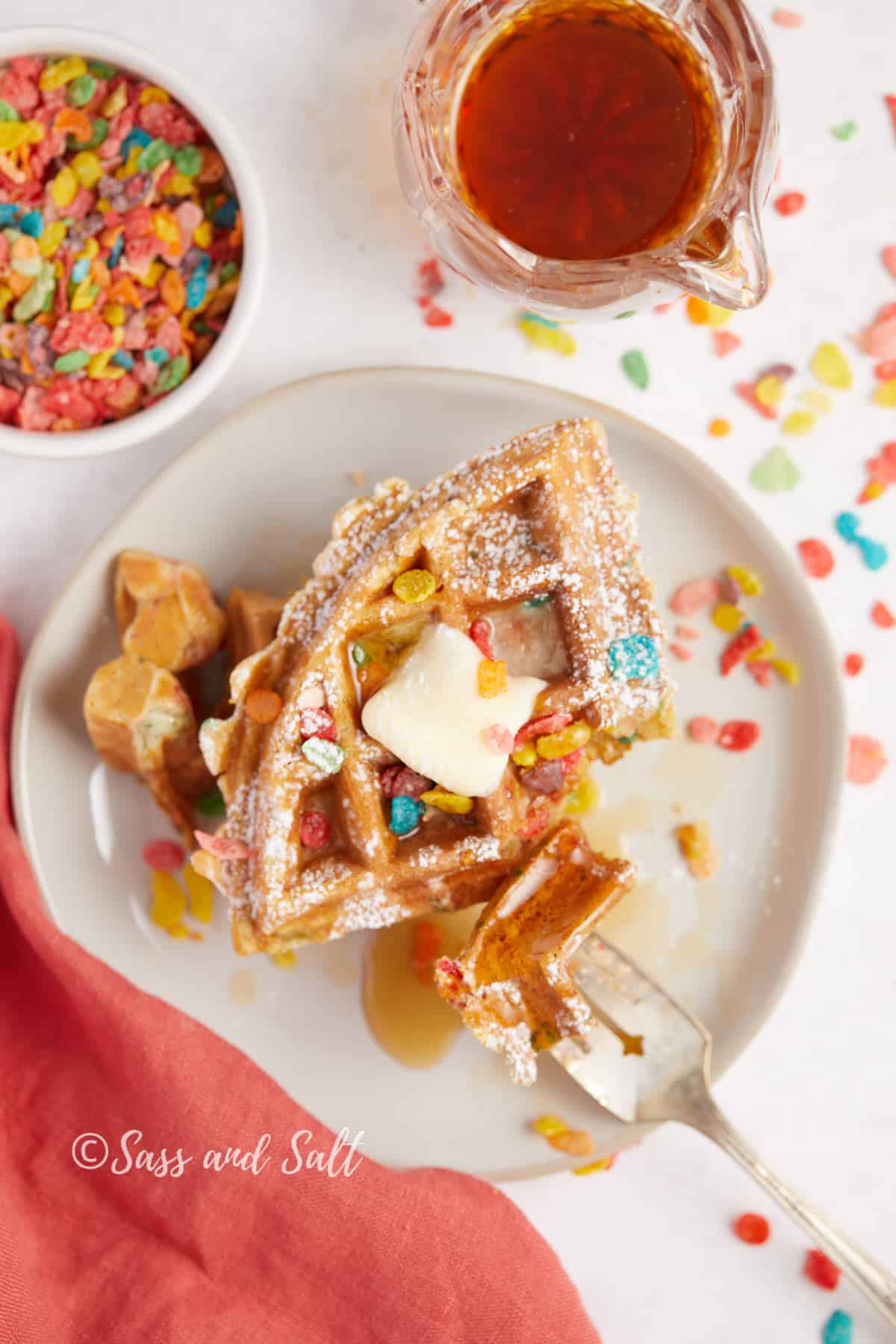 A decadent serving of Fruity Pebbles waffles topped with melting butter, drizzled with golden syrup, ready to be enjoyed alongside a cup of warm amber maple syrup.