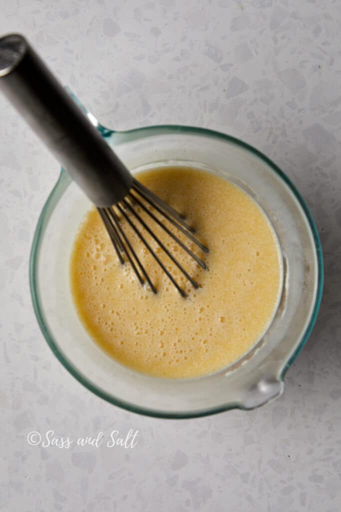 Overhead view of a whisk in a glass measuring cup with milk, melted butter, egg, and vanilla mixture mixed together sitting on a speckled counter top. 
