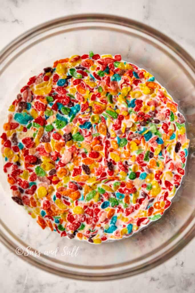 A clear glass bowl filled with milk and a generous amount of Fruity Pebbles cereal, fully covering the surface. The cereal's bright colors stand out vividly against the white milk, showcasing a mix of red, blue, green, yellow, and orange pieces. 