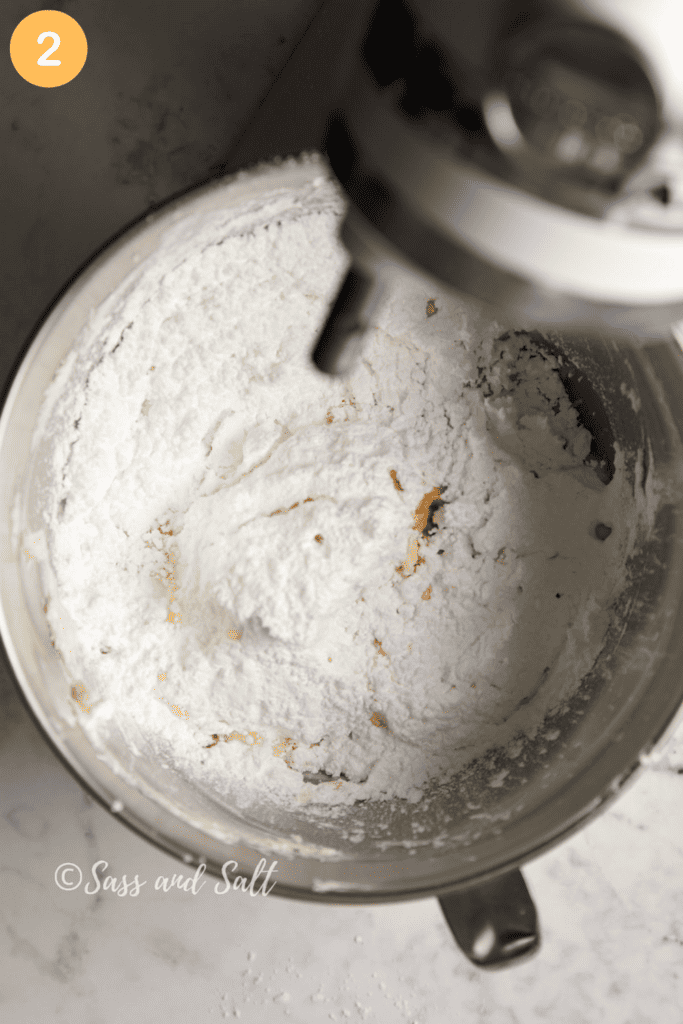 A mixing bowl filled with confectioner's sugar, butter, vanilla and milk mid-mix, showing the powdery texture beginning to combine, with the whisk attachment of a mixer in motion, all against a marble countertop backdrop.