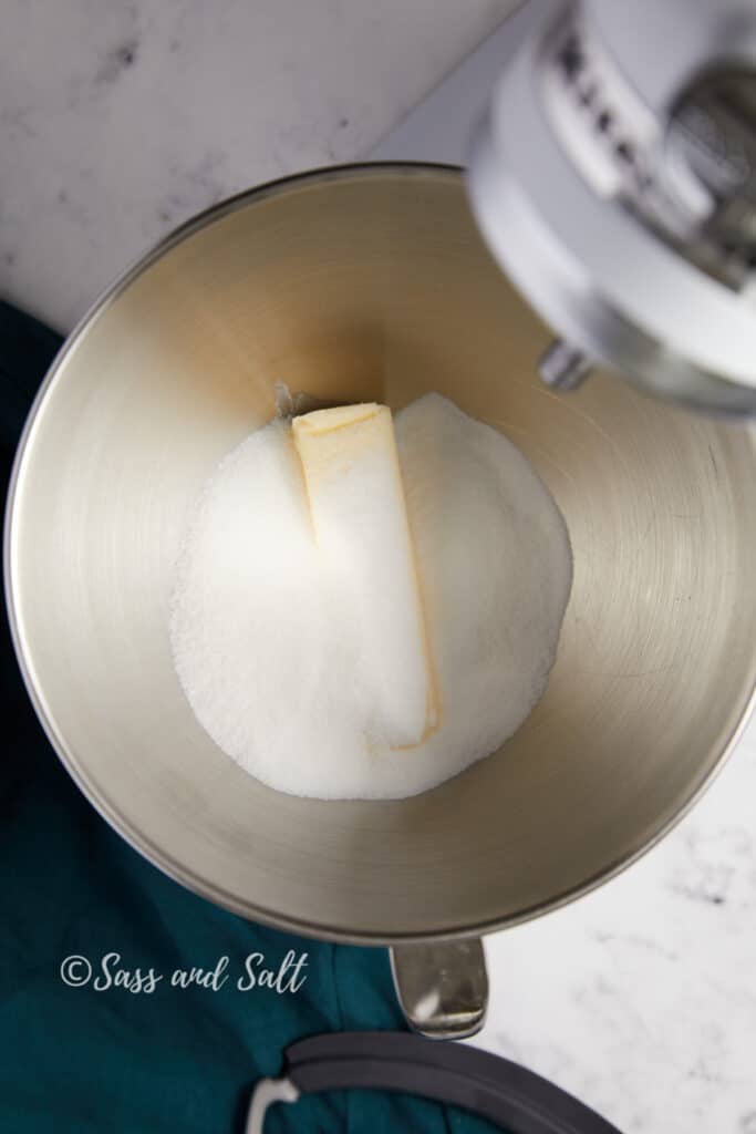 A stick of butter with a mound of sugar in a mixing bowl, positioned under a mixer's paddle attachment, all against a marble and teal background.