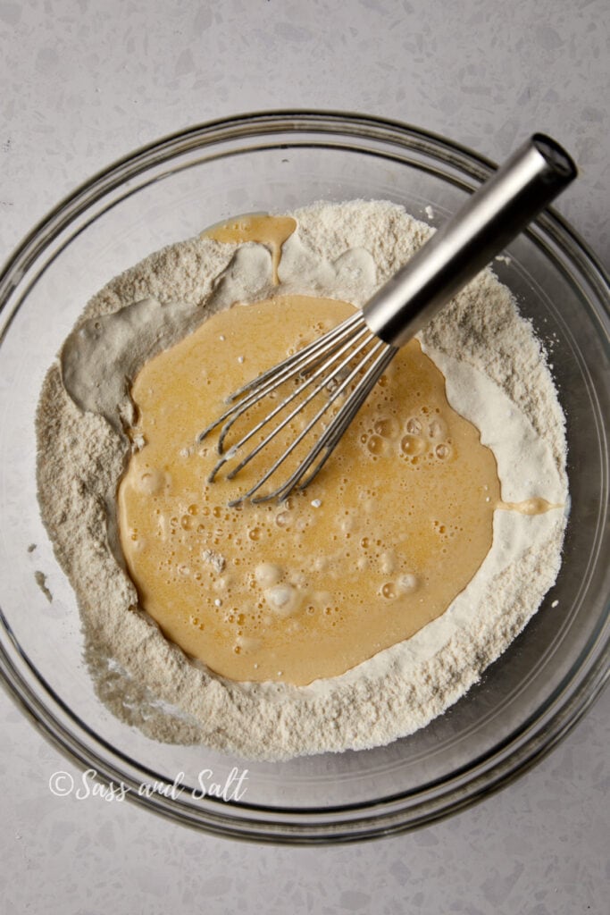 A clear glass bowl containing a mixture of dry flour with wet ingredients poured on top, starting the process of making waffle batter, with a whisk ready to combine them. 