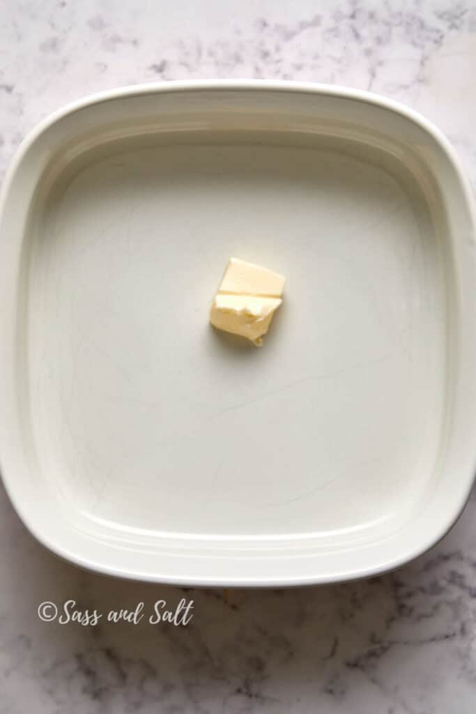 A single cube of butter in the center of a white rectangular baking dish placed on a marble surface.
