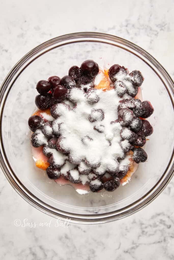 Top view of a in a glass bowl, featuring a layer of peach slices topped with cherries and generously dusted with sugar on a marble surface.