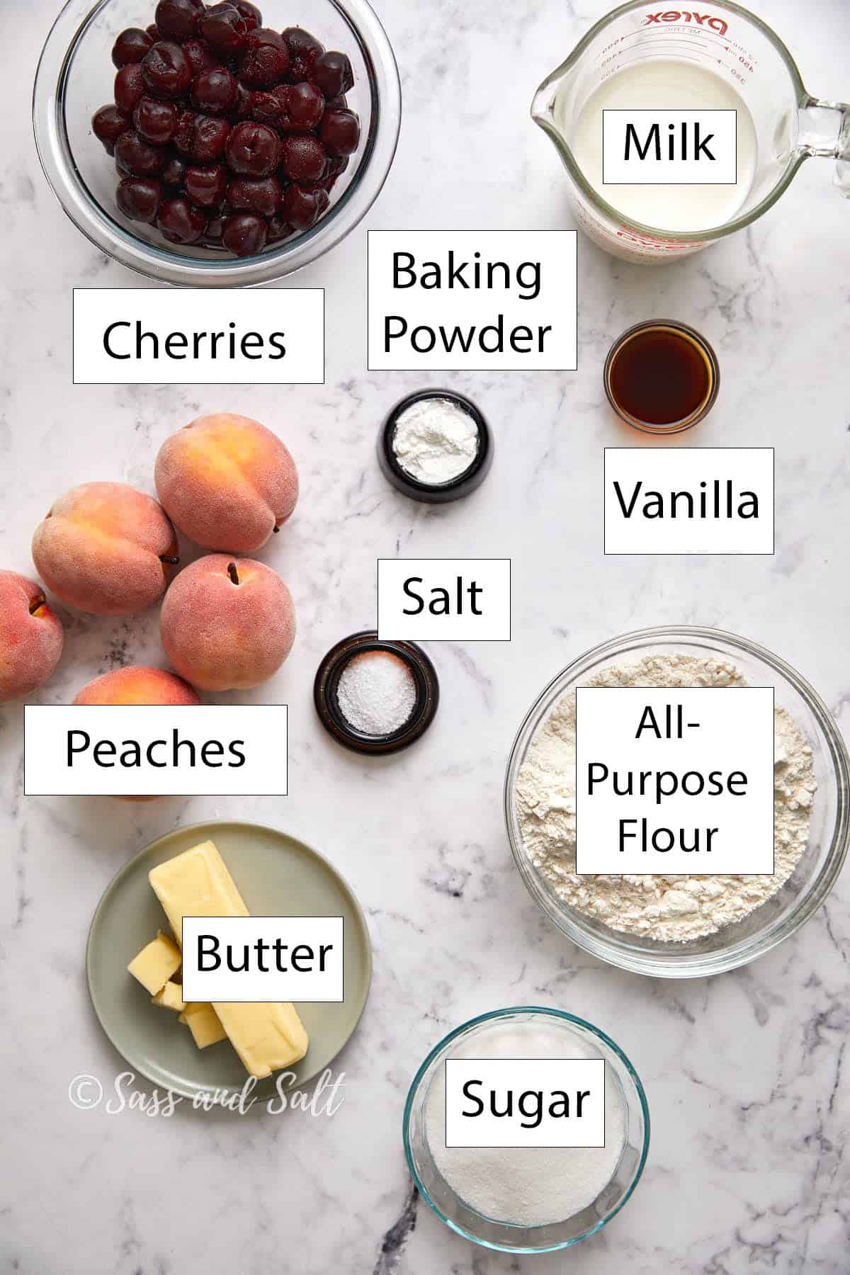 Flat lay of baking ingredients on a marble surface, labeled: cherries, peaches, butter, sugar, all-purpose flour, milk, vanilla, baking powder, and salt.