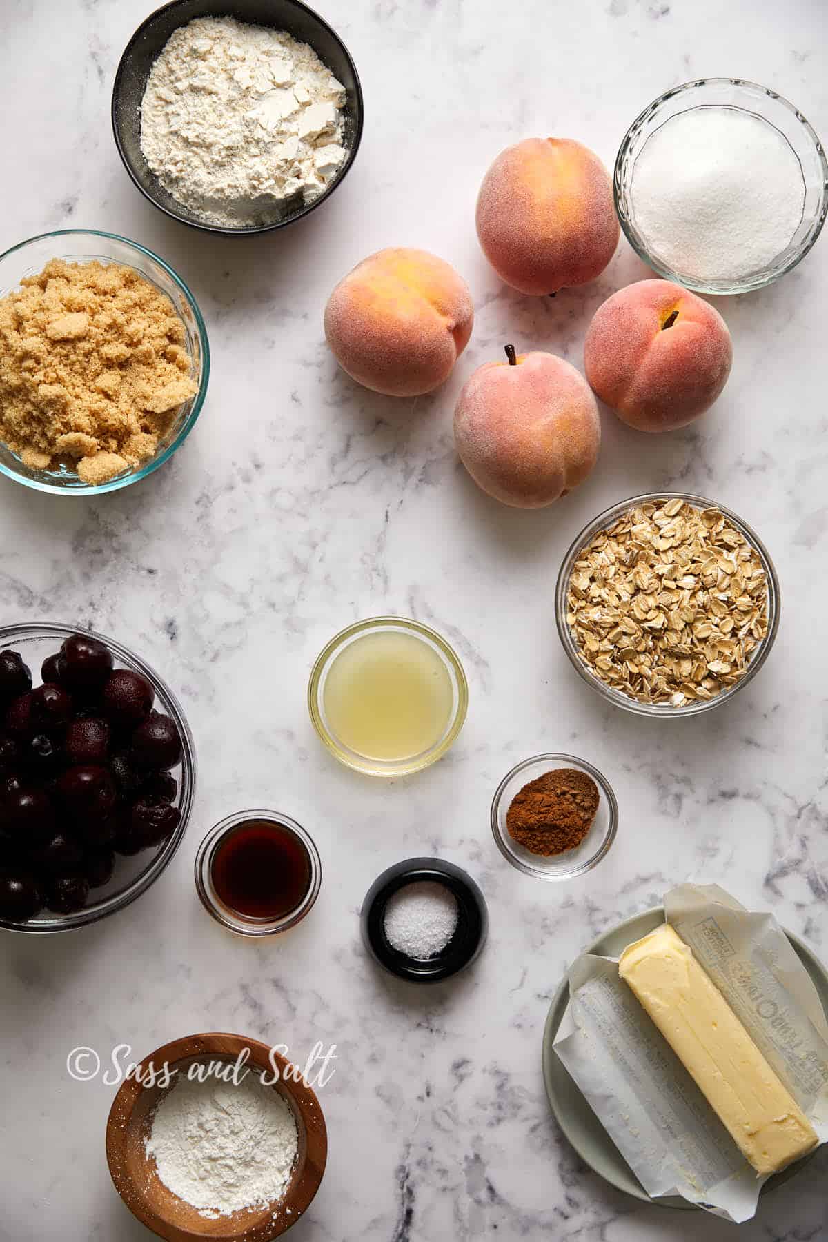 Various baking ingredients arranged on a marble surface, including flour, peaches, brown sugar, granulated sugar, oats, cherries,lemon juice, vanilla, corn starch, cinnamon, nutmeg, vanilla, and butter, viewed from above.