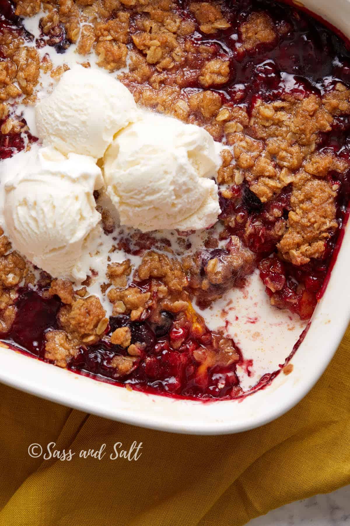 A freshly baked berry crumble topped with three scoops of vanilla ice cream, served in a white baking dish with vibrant red berry filling oozing from the sides.