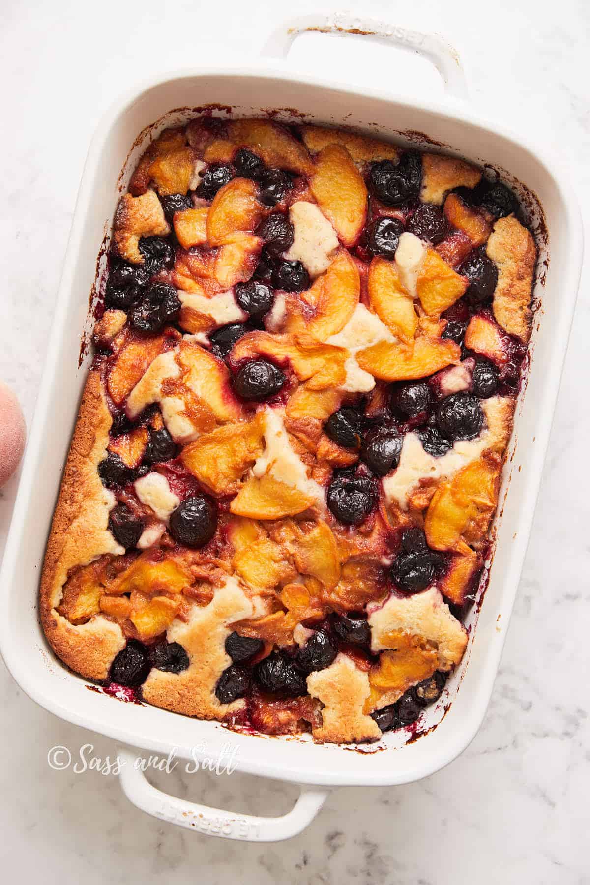 A freshly baked fruit cobbler in a white baking dish, featuring golden-brown crust with vibrant peaches and dark cherries visible.