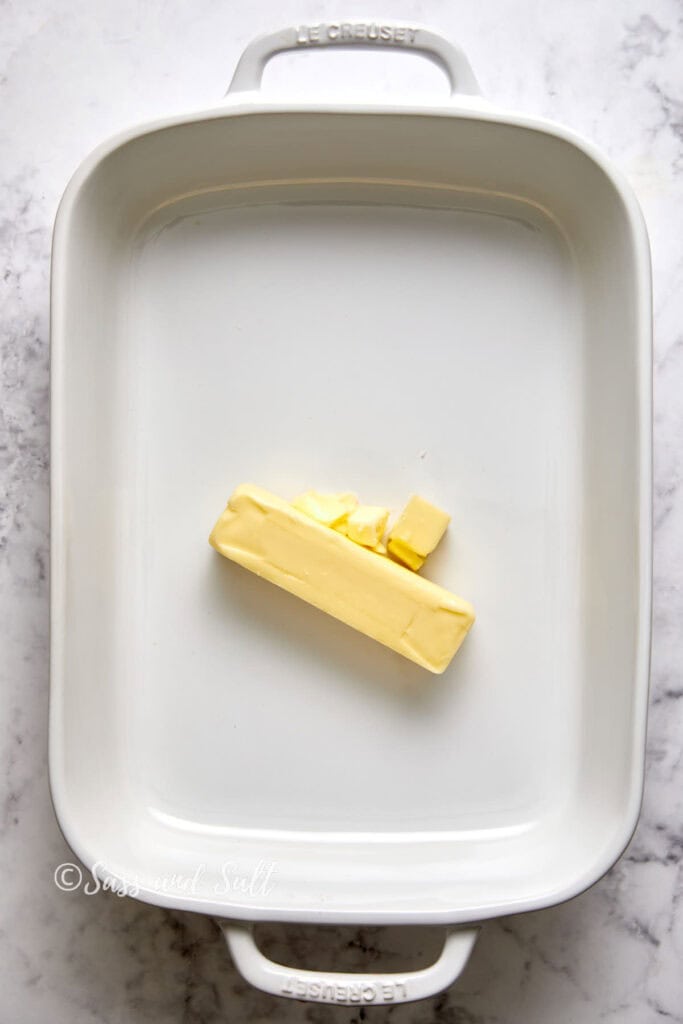 A stick of butter with a few pieces cut off, placed in the center of a white le creuset baking dish on a marble surface.