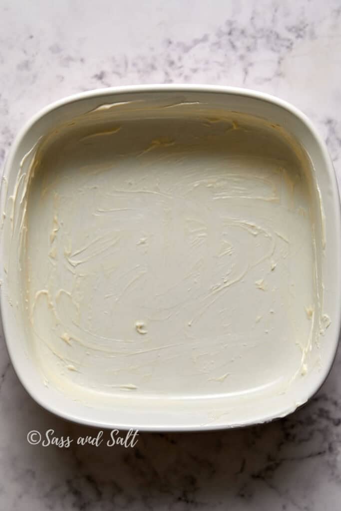 An image showing a rectangular container with butter creamed along its sides and bottom, on a marble surface. the texture is smooth with visible swirls. attribution: sass and salt.