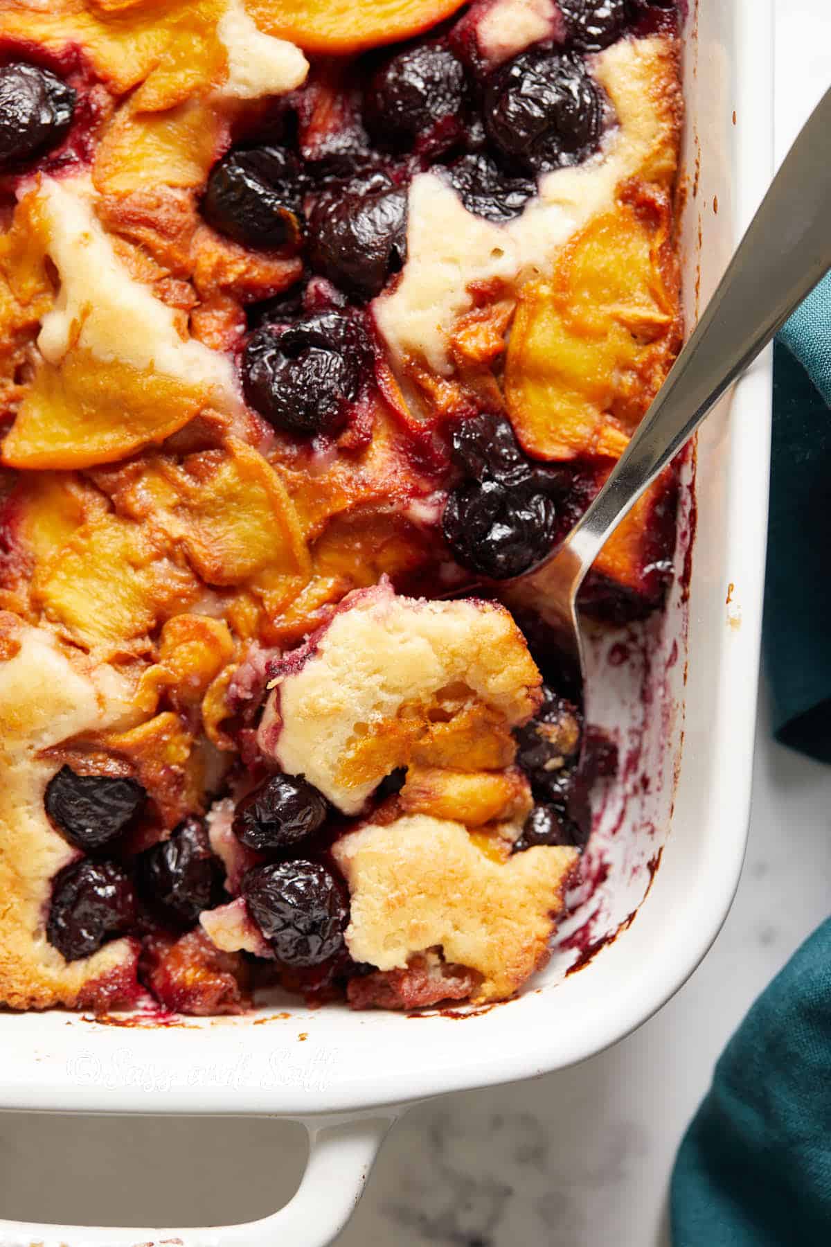 Close-up of a freshly baked fruit cobbler in a white dish, featuring vibrant cherries and peaches with a golden-brown crust, and a serving spoon on the side.