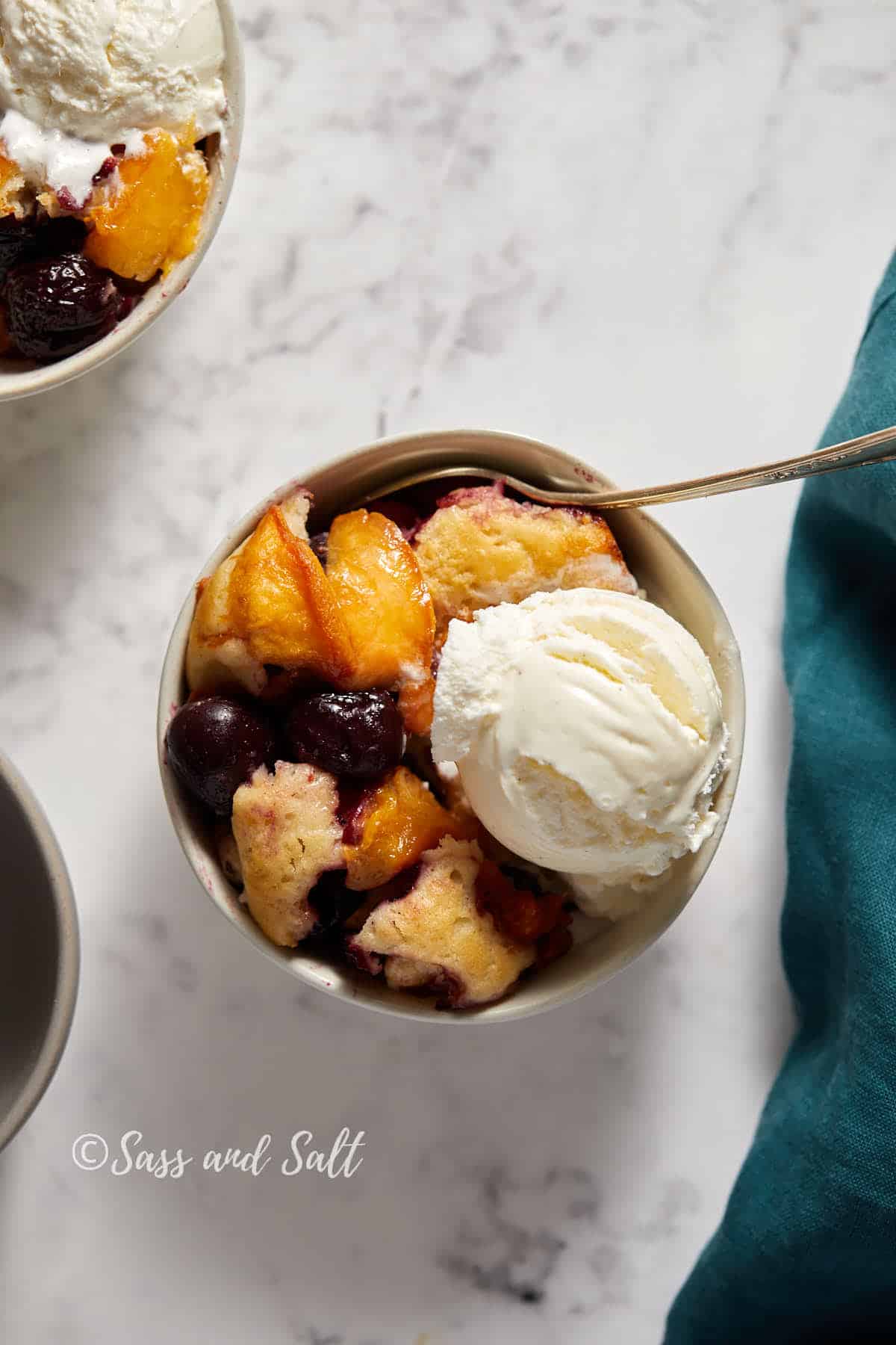A bowl of peach and cherry cobbler topped with a scoop of vanilla ice cream, captured from above on a marble surface with a teal napkin partially visible.