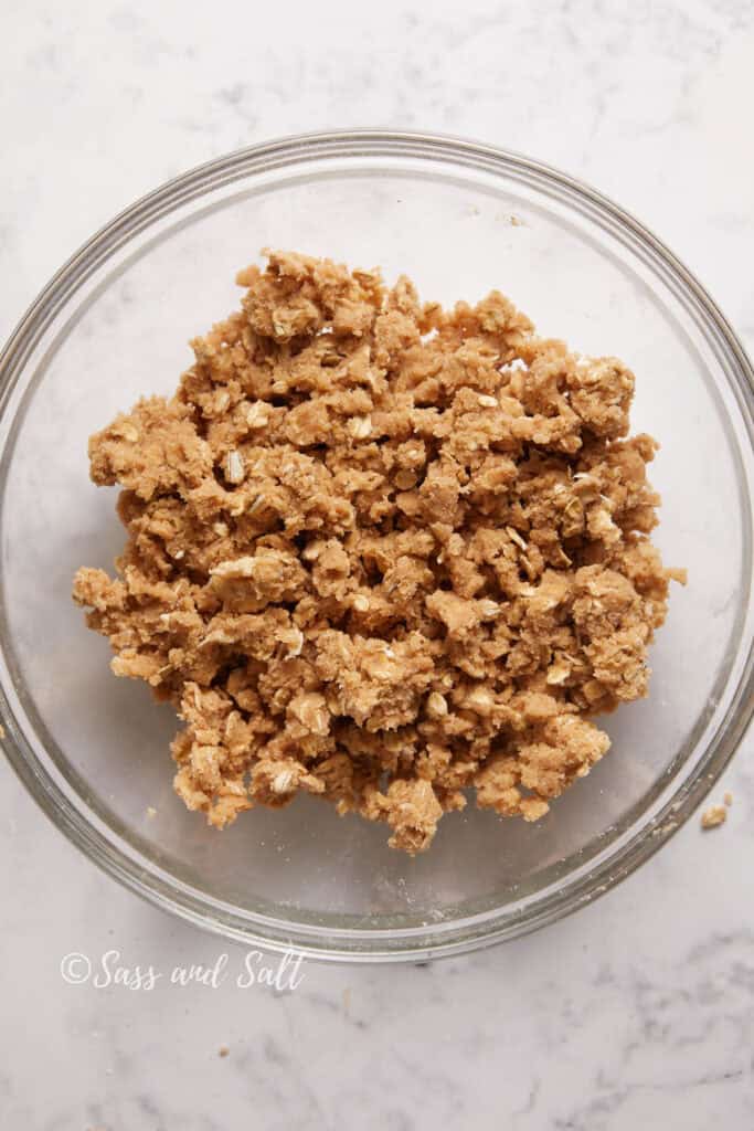 A clear glass bowl containing a pile of crumbled oatmeal  dough on a white marble countertop.