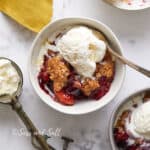A bowl of berry crumble topped with a scoop of vanilla ice cream, accompanied by a spoon, on a marbled surface with another similar dish nearby.