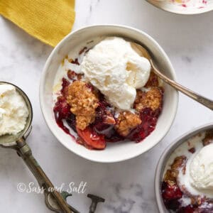 A bowl of berry crumble topped with a scoop of vanilla ice cream, accompanied by a spoon, on a marbled surface with another similar dish nearby.