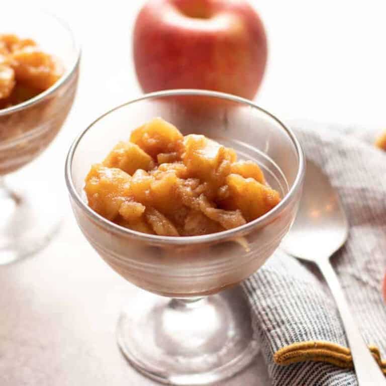 Two glass dessert bowls filled with homemade cinnamon chunky apple sauce are placed on a white surface, with a whole apple in the background. A striped cloth napkin and a spoon are also visible next to the bowls.