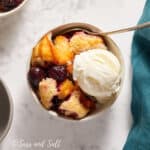 A bowl of peach cobbler topped with a scoop of vanilla ice cream, accompanied by fresh cherries, on a marble surface with a teal napkin.