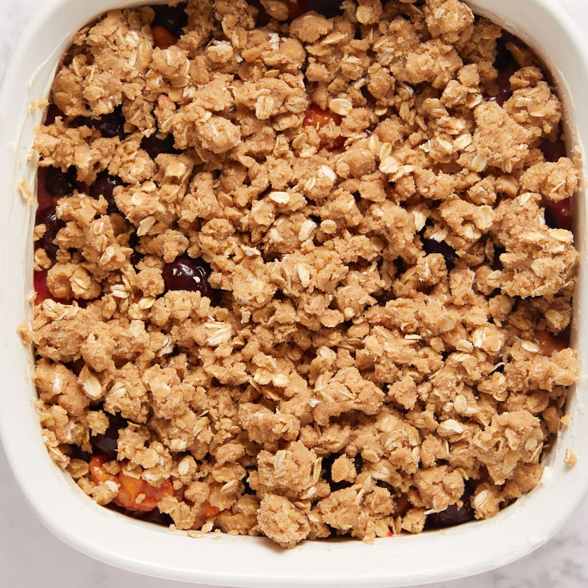 Oat streusel scattered over peaches and cherries in a white baking dish before baking.