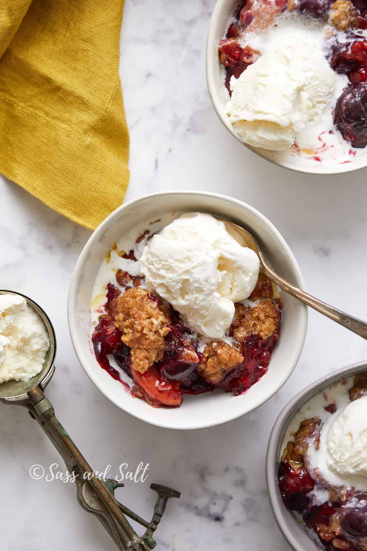 A bowl of peach and cherry crisp topped with a scoop of vanilla ice cream, beside a spoon and another dish of crisp, on a white marble surface with a yellow napkin.