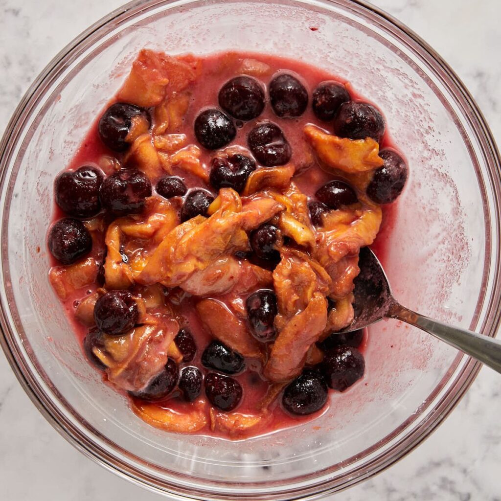 Sliced peaches and dark cherries tossed with sugar and cornstarch in a glass bowl, the filling for peach and cherry crisp.