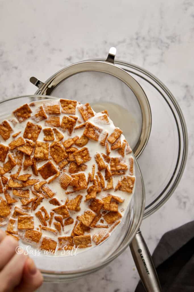 A bowl of Cinnamon Toast Crunch cereal being strained over a mesh strainer placed on another bowl. The cereal is soaked in milk, and the process appears to be straining the cereal-infused milk into the bowl below.