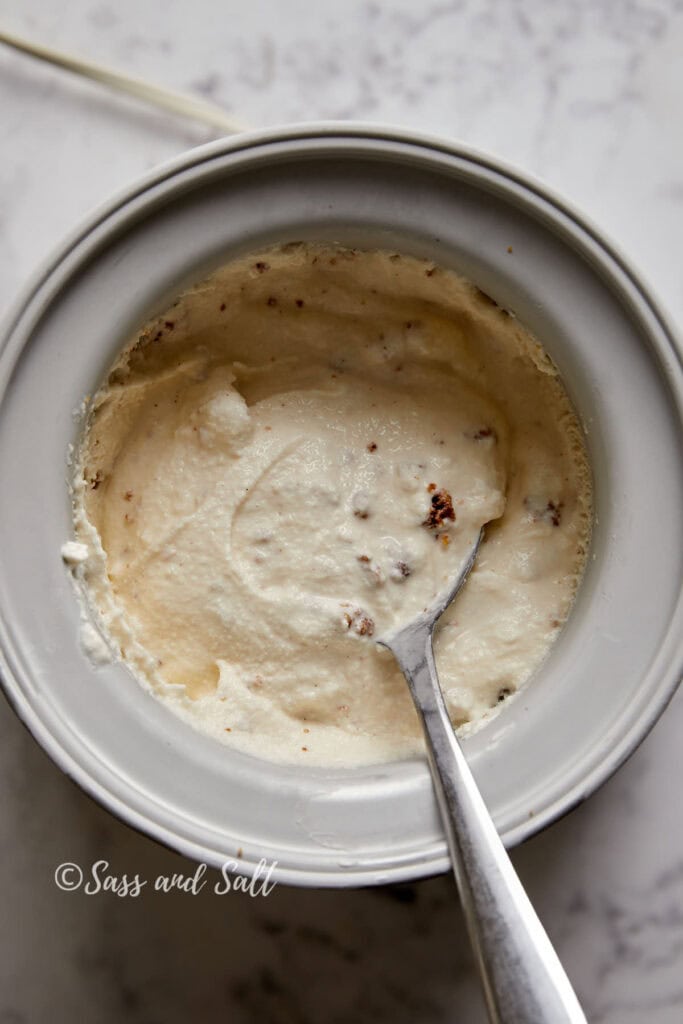 A cusinart ice cream freezer bowl filled with ice cream mixed with cinnamon toast crunch mix. A spoon sits inside the freezer bowl. The background is a white, slightly textured surface, and the text "Sass and Salt" appears in the bottom left corner.