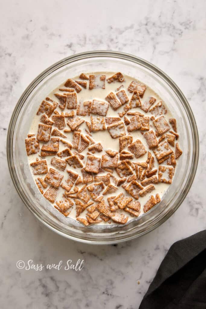 A glass bowl filled with milk and cinnamon toast crunch cereal mixed together is placed on a marble countertop. A small piece of dark fabric is visible in the lower right corner. "Sass and Salt" is written in the bottom left corner.