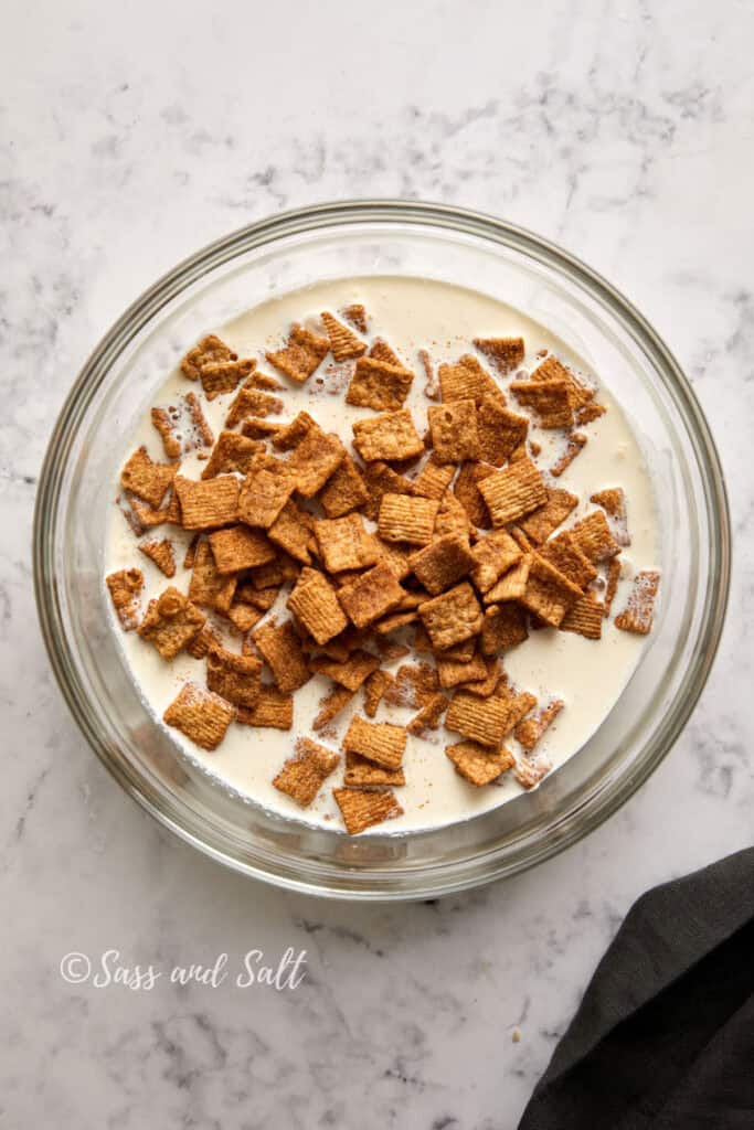 A glass bowl filled with cereal pieces and milk on a marble surface. The cereal pieces are golden-brown and square-shaped. There is a black cloth partially visible in the bottom-right corner. Text at the bottom-left corner reads "Sass and Salt.