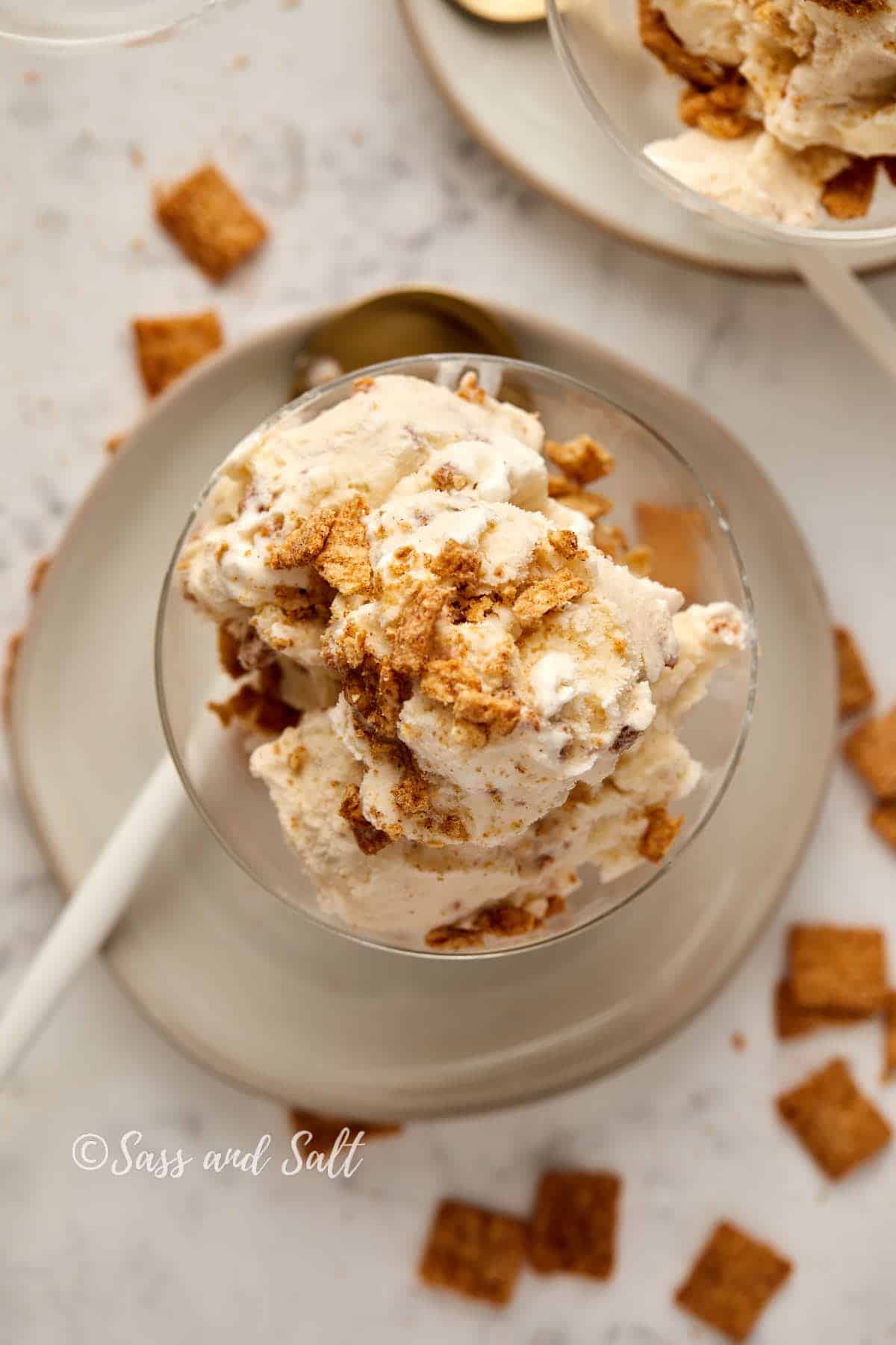 A glass bowl filled with creamy ice cream, topped with crunchy cereal pieces, sits on a white ceramic plate with a white spoon. The background includes a scattering of cereal pieces and partial views of other bowls on a marble surface.