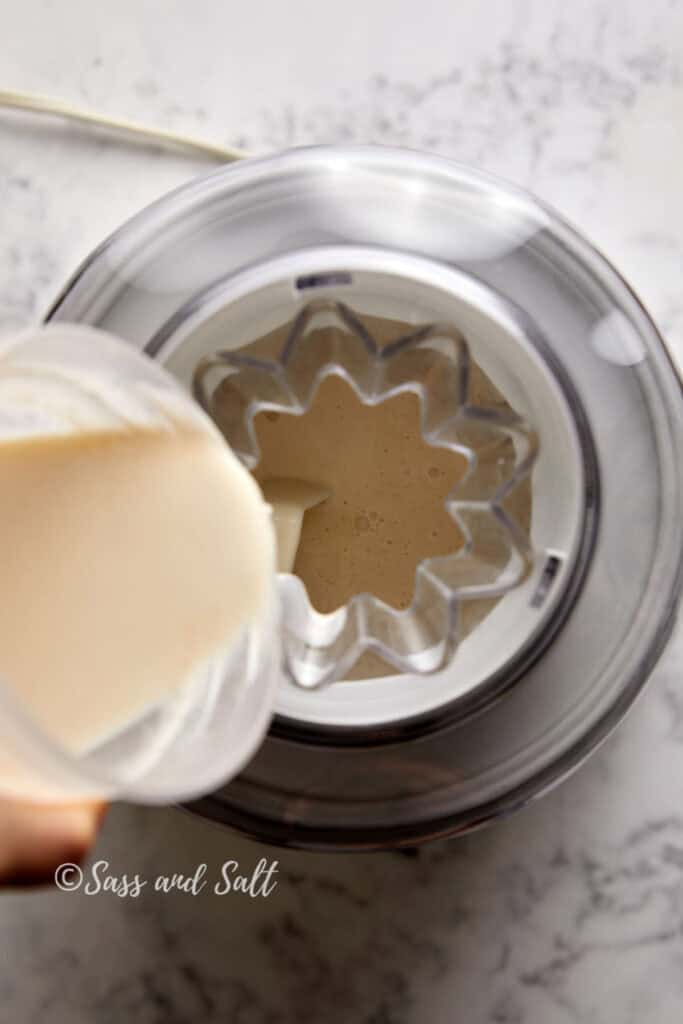 A top-down view of a person pouring a creamy mixture from a measuring cup into an ice cream maker on a marble countertop. "Sass and Salt" watermark is visible.