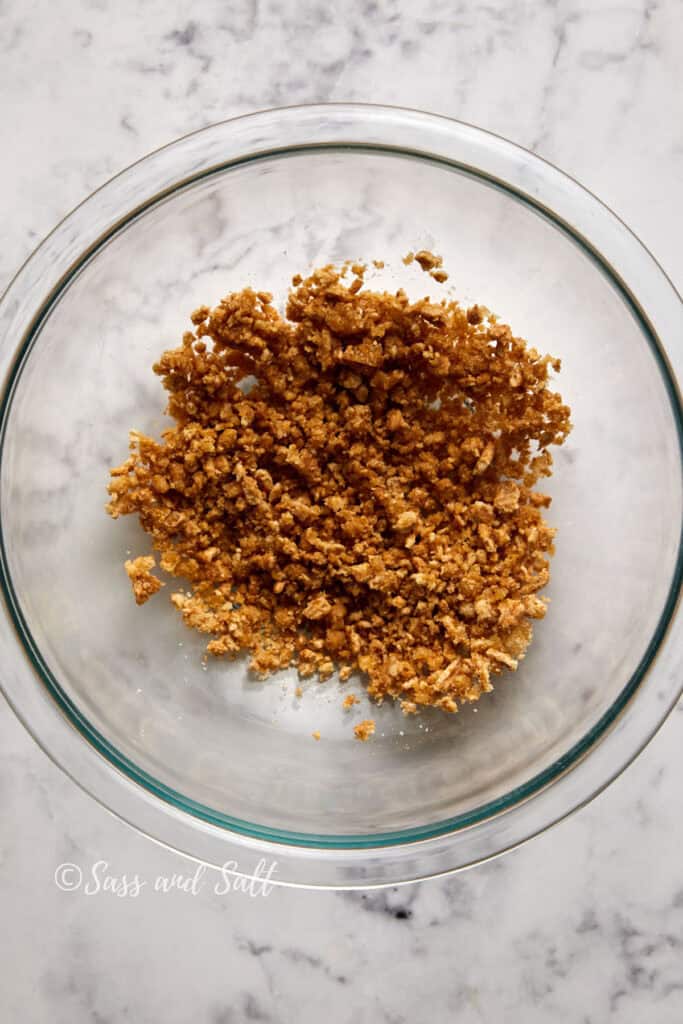 A glass bowl filled with crumbly, cinnamon toast crunch mixture sits on a marble surface. The mixture is golden brown in color and has a rough, uneven texture. The edge of the bowl features a "Sass and Salt" watermark.