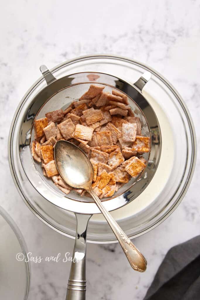 A strainer filled with cinnamon toast crunch cereal pieces sits atop a large metal bowl, ready for the next step in this amazing cinnamon toast crunch ice cream recipe. A spoon rests in the strainer, angled to the side. The setup is placed on a white marble surface.