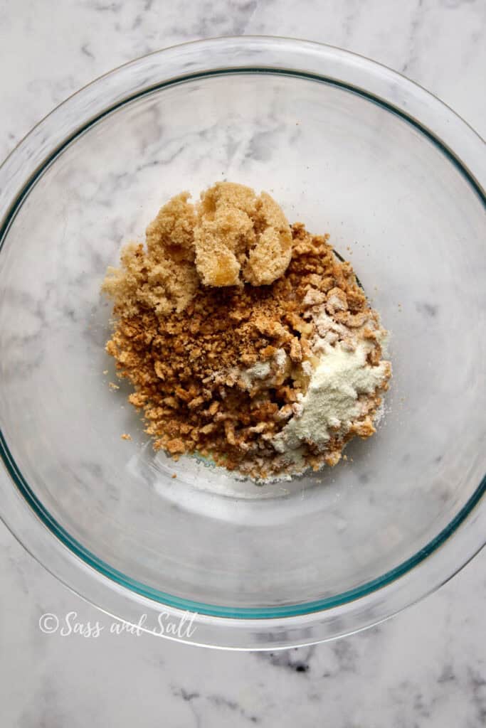 A clear glass bowl on a marble countertop contains a mixture of brown sugar, crushed cinnamon toast crunch, and milk powder. The ingredients are shown just before being mixed together for the cinnamon toast crunch mix for the ice cream base. There is text in the bottom left corner that reads "Sass and Salt.