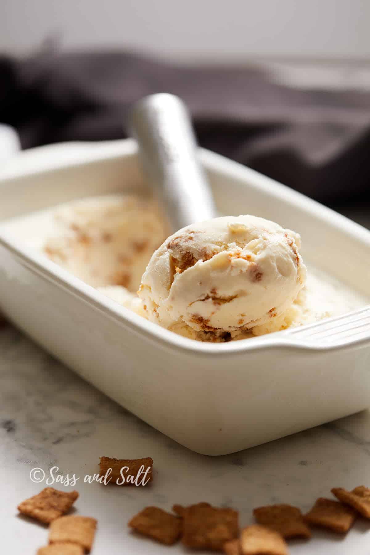 A close-up photo of a white rectangular dish filled with creamy ice cream, featuring a metal ice cream scoop resting on top with a scoop of ice cream. The dish is placed on a marble surface next to several small, square pieces of cereal.