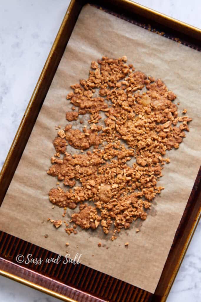 A baking tray lines with parchment paper and holds a spread of golden brown, crunchy clusters of cinnamon toast crunch mix. The backdrop includes a marble countertop.