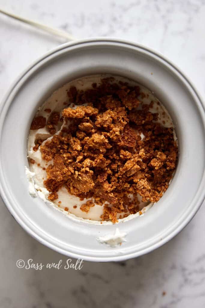 A close-up of a white ice cream freezer bowl filled with ice cream topped with cinnamon toast crunch mix. The bowl is on a marble surface.