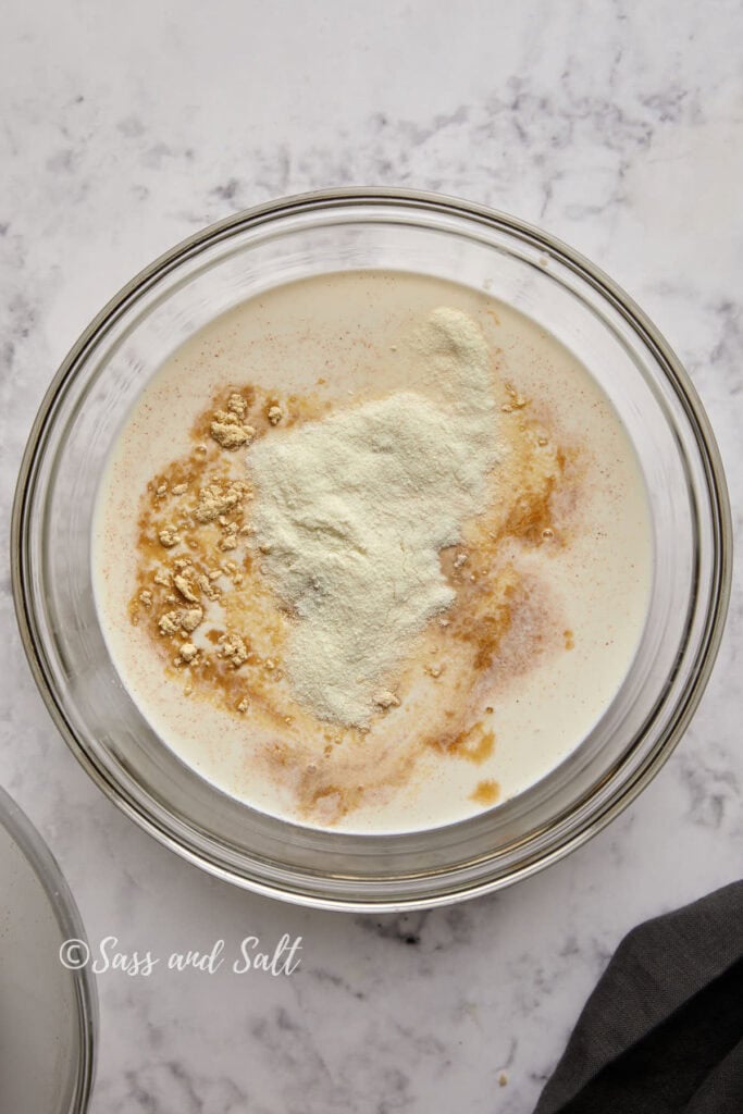 A clear glass bowl sits on a marble countertop, containing a mixture of milk and coarse granules, likely oatmeal, with some powder on top. A black cloth is partially visible in the bottom right corner. The text "Sass and Salt" is printed on the left side of the bowl, evoking thoughts of a cinnamon toast crunch ice cream recipe.