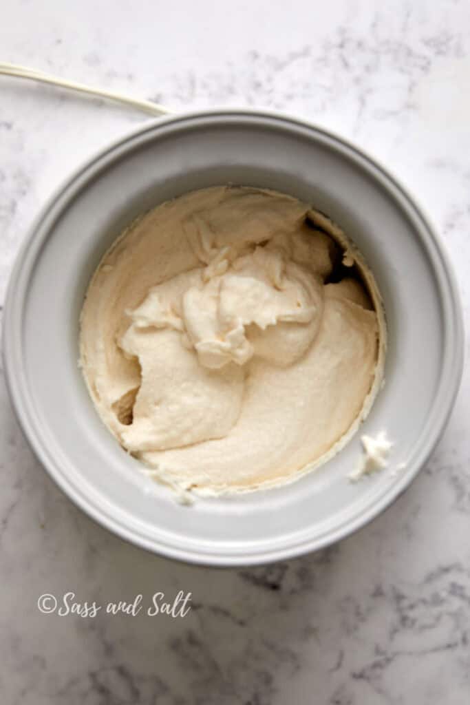 Overhead view of a metallic ice cream maker bowl with a creamy cinnamon toast crunch ice cream mixture inside. The bowl is set on a white marble countertop. Text at the bottom left corner reads "© Sass and Salt.