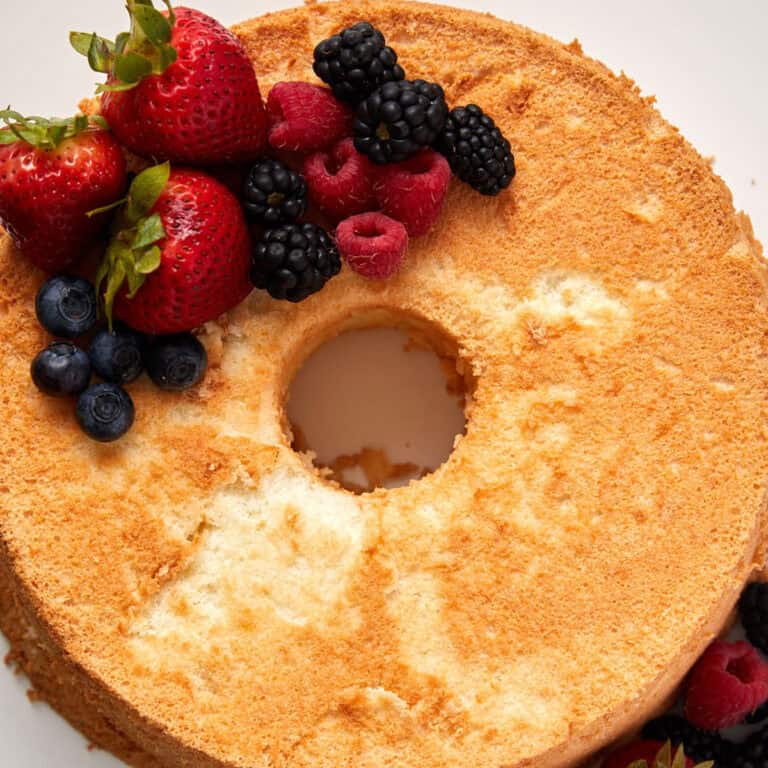 A golden brown, fluffy angel food cake with a hole in the center is garnished with fresh strawberries, raspberries, blackberries, and blueberries arranged on the top left section. The cake's airy texture is visible with slight cracking on the surface.