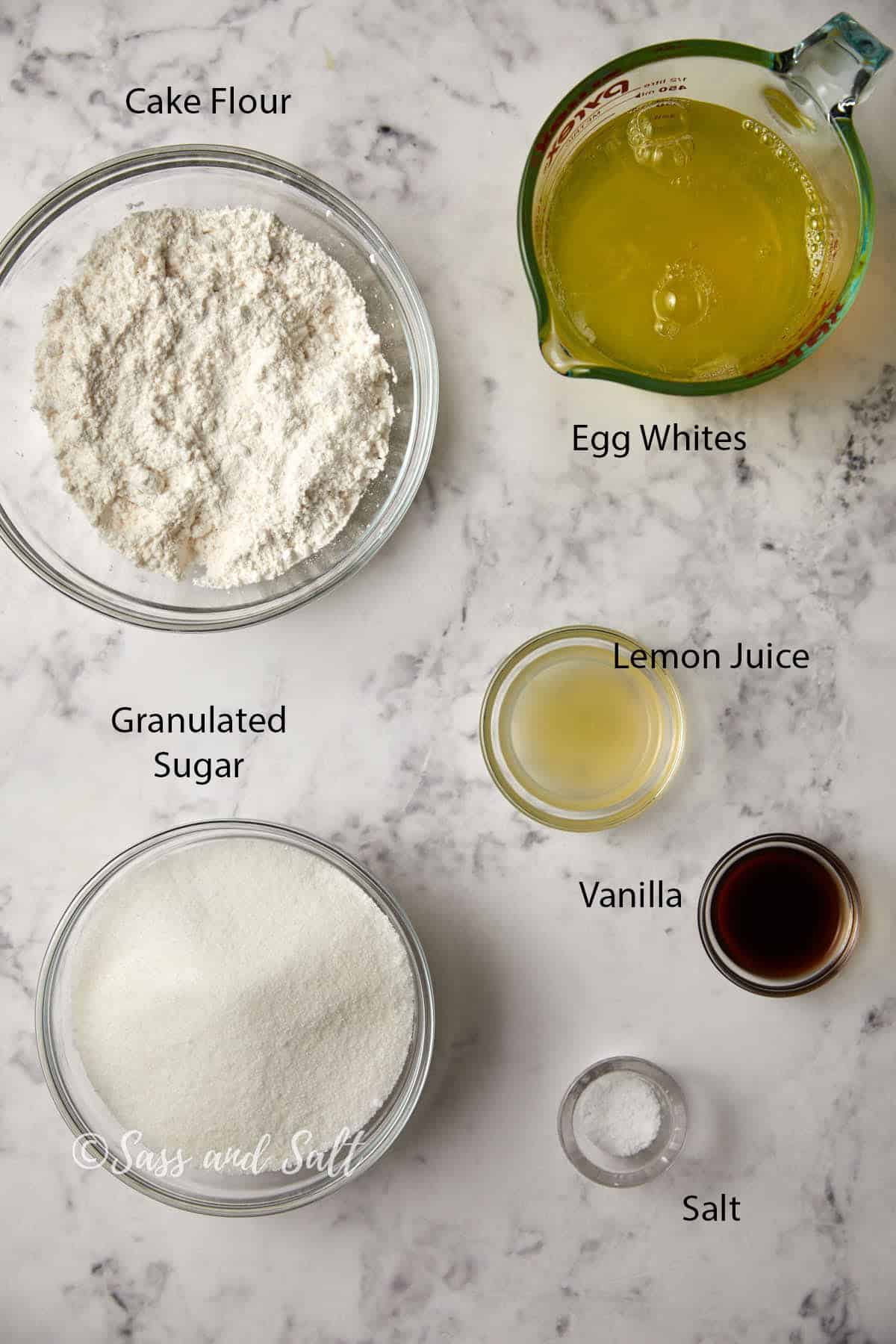 A marble countertop with ingredients labeled for baking. Ingredients include a bowl of cake flour, a measuring cup with egg whites, a bowl of granulated sugar, a small bowl of lemon juice, a small bowl of vanilla extract, and a pinch of salt.