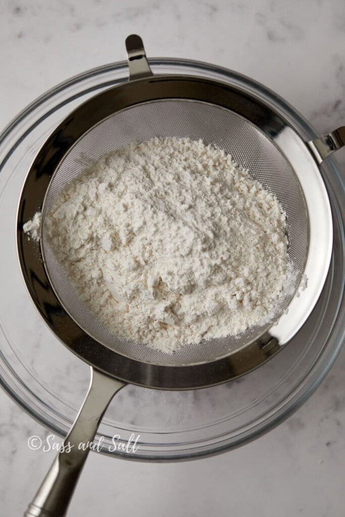 A metal sieve containing flour is placed over a clear glass bowl on a marble surface. The sieve shows some flour along with small chunks, indicating it is being sifted. The words "Sass and Salt" are visible in the bottom left corner of the image.